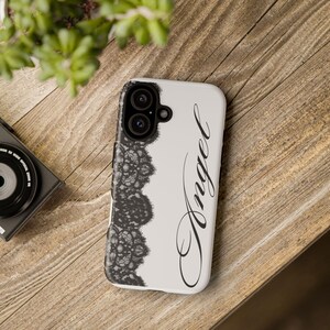 May include: A white phone case with a black lace design on the left side and the word "Angel" in cursive script. The phone case has a dual-lens camera cutout. The case is on a wooden surface.