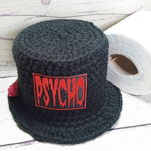 May include: A black crocheted toilet roll holder featuring a red "PSYCHO" patch. The holder is cylindrical with a small brim and a small red flower. A roll of white toilet paper is visible in the background.