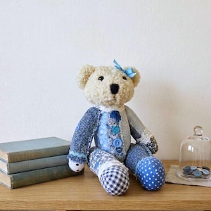 Puede incluir: Un oso de peluche hecho a mano con una cabeza color crema y un lazo azul. El cuerpo del oso está hecho de mezclilla de retazos en varios patrones azules. Está sentado sobre una superficie de madera junto a una pila de libros y una campana de cristal.