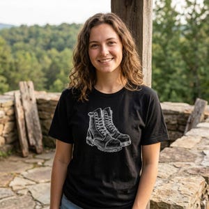 May include: A black short-sleeved t-shirt featuring a white graphic illustration of a pair of lace-up work boots centred on the chest. The design depicts the boots with detailed stitching, eyelets, and textured soles in a sketch-like style.