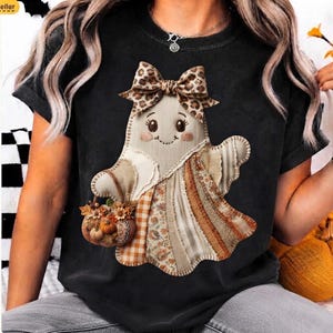 Puede incluir: Una camiseta negra de manga corta con el gráfico de un fantasma que lleva un lazo con estampado de leopardo. El fantasma tiene un diseño de retazos en tonos beige, marrón y naranja y sostiene una pequeña cesta llena de calabazas otoñales.