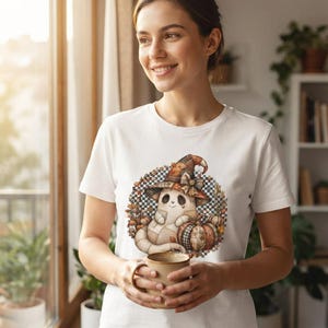 以下が含まれることがあります： パッチワーク風の魔女の帽子をかぶり、カボチャの横に座っているゴーストのグラフィックプリントが施された白い半袖Tシャツ。デザインは、秋の葉と白黒のチェック柄の円形の縁取りで囲まれています。