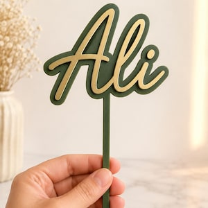 Pode incluir: Um topo de bolo verde com o nome "Ali" em escrita dourada. O topo é segurado por uma haste verde, e o design apresenta um efeito em camadas. O fundo é de uma cor suave e neutra, com um vaso de flores secas visível.