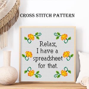 May include: A square cross stitch pattern framed in light wood featuring the black text "Relax, I have a spreadsheet for that." surrounded by six small yellow roses with green leaves arranged in a symmetrical grid on a white grid background.