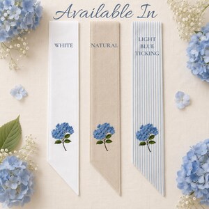 Puede incluir: Tres tiras rectangulares de tela etiquetadas como Available In, en opciones blanco, natural y rayas azul claro. Cada tira muestra un bordado de flor de hortensia azul en la parte inferior, rodeado de hortensias azules y flores de paniculata.
