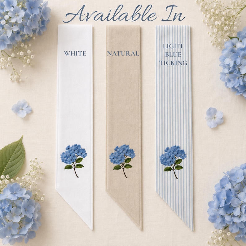 Puede incluir: Tres tiras rectangulares de tela etiquetadas como Available In, en opciones blanco, natural y rayas azul claro. Cada tira muestra un bordado de flor de hortensia azul en la parte inferior, rodeado de hortensias azules y flores de paniculata.