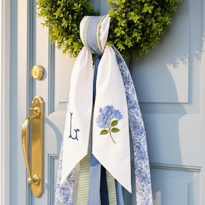 Puede incluir: Una corona de boj verde adornada con una cinta decorativa con extremos de tela blanca, uno bordado con una flor de hortensia azul y el otro con una letra monograma L azul marino, acentuada por cintas de tela con motivos florales y rayas.