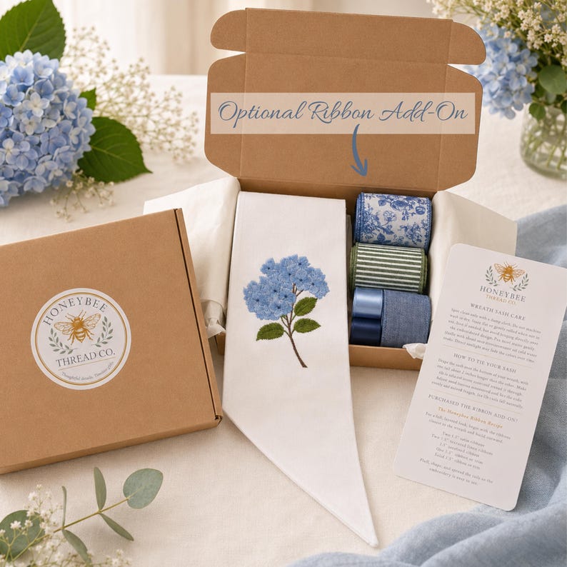 Puede incluir: Una banda de tela blanca con una flor de hortensia azul bordada, presentada en una caja de cart&oacute;n abierta con tres cintas enrolladas, una pegatina de Honeybee Thread Co. en la tapa y una tarjeta con instrucciones de cuidado y uso.