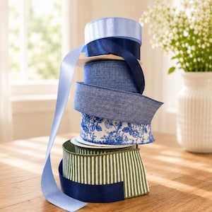 Puede incluir: Una pila vertical de seis carretes de cinta con diversas texturas y patrones. La colecci&oacute;n incluye azul claro s&oacute;lido, azul marino oscuro, azul mezclilla texturizado, una cinta blanca con estampados florales azules y una cinta a rayas verdes y blancas.