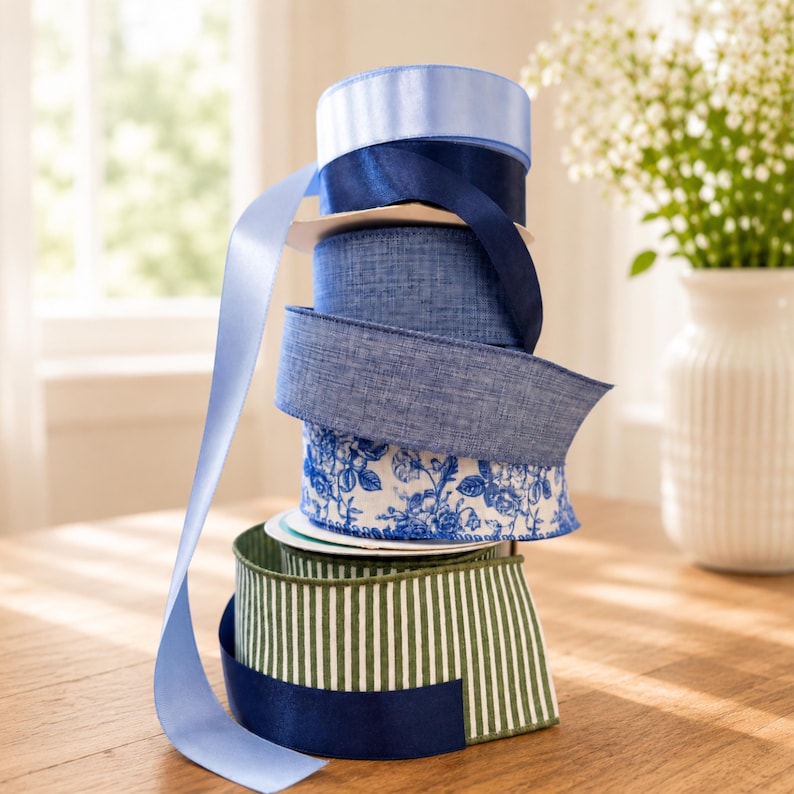 Puede incluir: Una pila vertical de seis carretes de cinta con diversas texturas y patrones. La colecci&oacute;n incluye azul claro s&oacute;lido, azul marino oscuro, azul mezclilla texturizado, una cinta blanca con estampados florales azules y una cinta a rayas verdes y blancas.