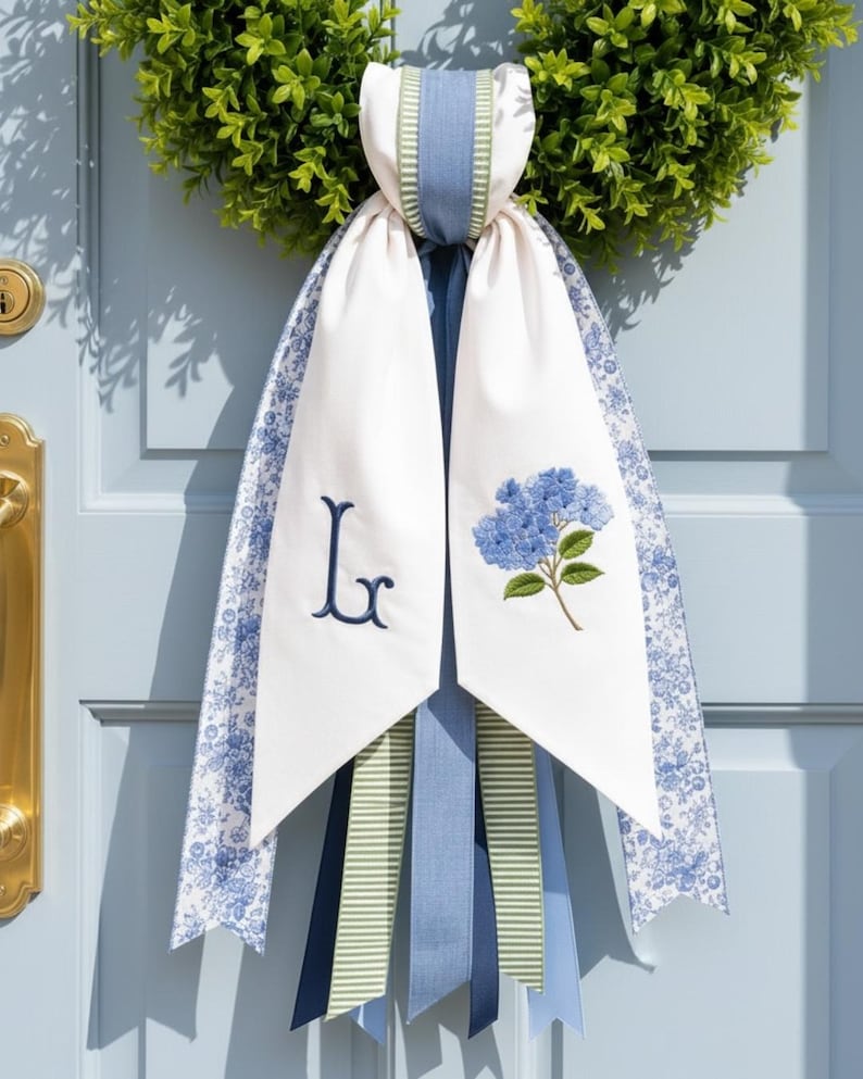 Puede incluir: Un colgador de puerta decorativo de tela con una base de corona de boj verde y cintas largas superpuestas. Las cintas incluyen paneles blancos con una letra monograma L bordada en azul marino y una flor de hortensia azul, adem&aacute;s de patrones florales y de rayas azules.