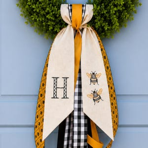 May include: A decorative wreath bow with a green wreath base. The bow features a cream-colored banner with a black embroidered "H" and two embroidered bees. The bow has black and white checkered ribbon, and yellow and black patterned ribbon.