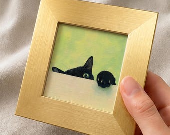 May include: A framed artwork featuring a black cat peeking over a white surface against a light green background. The cat's ear, eye, and paw are visible. The frame is gold-colored.