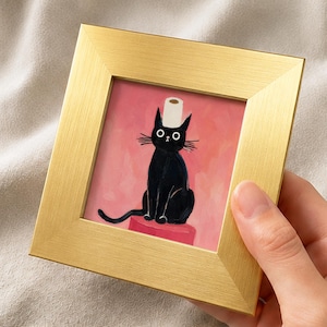 Pode incluir: Uma impressão artística quadrada emoldurada apresentando um gato preto de olhos arregalados sentado em um pedestal vermelho. O gato tem um rolo de papel higiênico branco equilibrado na cabeça. O fundo é rosa. A moldura tem acabamento dourado.