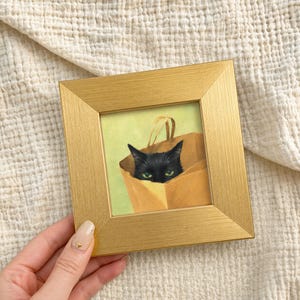Könnte beinhalten: Gerahmtes Gemälde einer schwarzen Katze, die aus einer braunen Papiertüte schaut. Die Katze hat grüne Augen und ist vor einem hellgrünen Hintergrund platziert. Der Rahmen ist goldfarben.