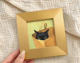 May include: A framed painting depicts a black cat peeking out of a brown paper bag. The cat has green eyes and is set against a light green background. The frame is gold-colored.