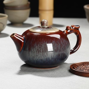 Puede incluir: Tetera de cerámica vidriada en tonos rojo oscuro y azul. La tetera tiene un cuerpo redondeado, un asa curva y un pico. La tapa está rematada con un pequeño pomo. Se ve un posavasos de madera. Juego de té.