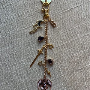 May include: A gold-colored keychain with various charms, including a crescent moon, stars, a sword, a dragon, and a lightning bolt. The charms are attached to a gold chain and a heart-shaped clasp.