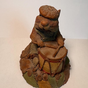 May include: A detailed figurine of a squirrel playing a drum. The squirrel wears a brown hat and overalls, and the drum is tan with red accents. The base is decorated with leaves and acorns, creating a whimsical, nature-inspired scene.
