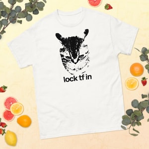 Peut inclure: T-shirt blanc avec un motif noir et blanc d'un visage de chat et le texte "lock tf in" en dessous. Le motif du chat est détaillé et texturé. Vêtement de style décontracté.