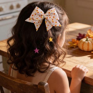 Peut inclure: Un nœud pour cheveux à fond blanc avec un motif de petits bonbons en forme de maïs orange, jaune et blanc, agrémenté de deux petites pinces à cheveux en forme d'étoile jaune et rose, fixées dans des cheveux bruns ondulés.