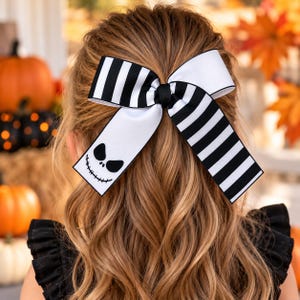 May include: A hair bow featuring a black and white striped pattern on one side and a solid white section on the other. The white section displays a black graphic of a smiling face with stitched mouth details and large, almond-shaped eyes.