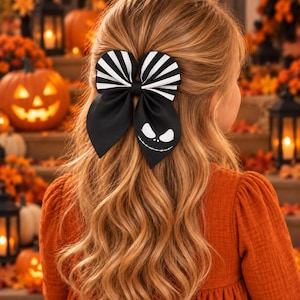 May include: A hair bow featuring a black and white striped top section and a solid black bottom section. The bottom right tail of the bow displays a white graphic of a character face with large oval eyes and a wide, stitched-line mouth design.