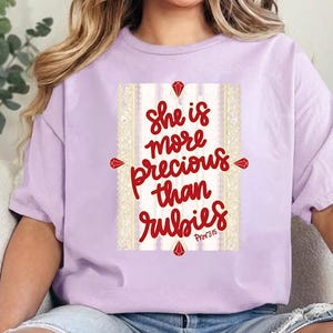 Puede incluir: Camiseta lila claro con la frase "She is more precious than rubies" en letras rojas. El texto está dentro de un marco decorativo con detalles de gemas rojas. Prenda de vestir informal.
