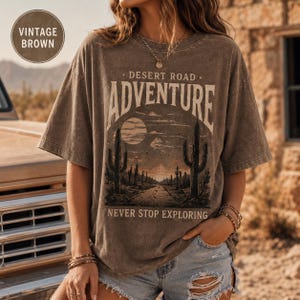 Puede incluir: Una camiseta marrón de manga corta con un estampado gráfico de un paisaje desértico con cactus, una carretera y un sol. El texto dice "DESERT ROAD ADVENTURE" y "NEVER STOP EXPLORING" en letras blancas. La camiseta tiene un aspecto desgastado.
