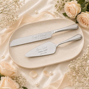Puede incluir: Un juego de cuchillo y servidor de pastel plateado sobre un plato ovalado crema. El cuchillo está grabado con "Emily & Daniel" y la fecha "June 18, 2026". El servidor dice "Love is Sweet" y la misma fecha. Flores.