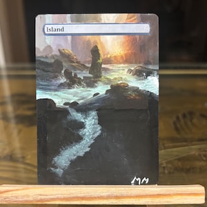 May include: A hand-painted card depicting an island scene with a rocky shoreline and a bright sunset. The word "Island" is at the top. The card is displayed on a small wooden easel.