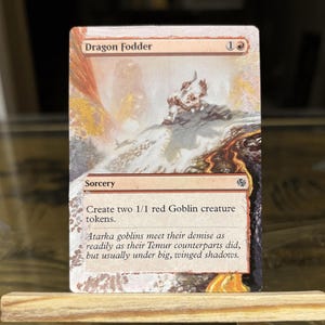 May include: A Magic: The Gathering card titled "Dragon Fodder" with a fantasy landscape illustration. The card text reads "Create two 1/1 red Goblin creature tokens." The card is displayed on a small wooden easel.