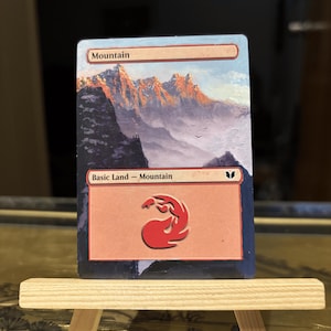 May include: A Magic: The Gathering card titled "Mountain" displayed on a small wooden easel. The card features a landscape painting of a mountain range with a red symbol at the bottom. The sky is light blue.