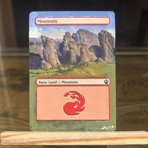 May include: A hand-painted card featuring a mountain landscape with a red symbol. The card has the word "Mountain" at the top and "Basic Land - Mountain" at the bottom. The artwork depicts a rocky mountain range under a blue sky.