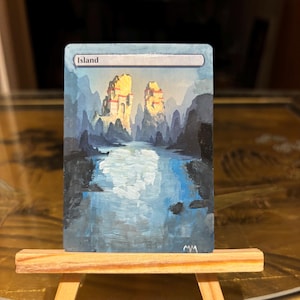 May include: Hand-painted artwork of an island scene. The artwork features a blue and white river flowing between dark cliffs, with two golden structures in the background. The word "Island" is at the top. The artwork is displayed on a small wooden easel.