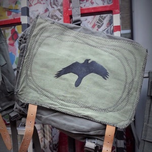 May include: A green canvas rucksack with brown leather straps and a black silhouette of a bird on the front. The rucksack has a zipped pocket on the front and a large main compartment.
