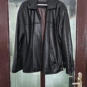 May include: A black leather jacket with a collared neckline and a zipper closure. The jacket has two front pockets and is hanging on a white hanger. The interior lining is visible.