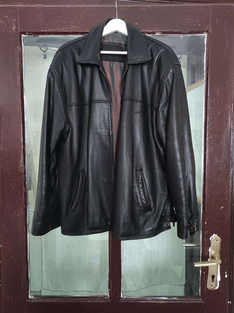 May include: A black leather jacket with a collared neckline and a zipper closure. The jacket has two front pockets and is hanging on a white hanger. The interior lining is visible.