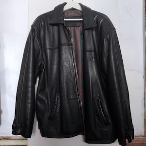 May include: A black leather jacket with a collared neckline and a zipper closure. The jacket has two front pockets and is hanging on a white hanger. The interior lining is visible, featuring a striped pattern.