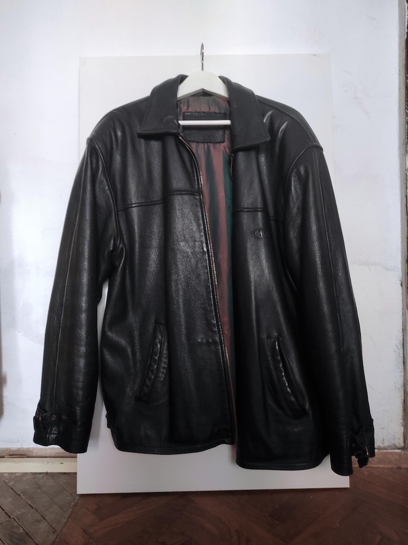 May include: A black leather jacket with a collared neckline and a zipper closure. The jacket has two front pockets and is hanging on a white hanger. The interior lining is visible, featuring a striped pattern.