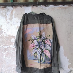 Può includere: Una camicia a maniche lunghe verde oliva con un grande pannello rettangolare a punto croce sul retro. Il pannello mostra un colorato bouquet di fiori viola, rosa e bianchi in un vaso di vetro, con la scritta Walle Rödel stampata in basso.