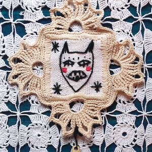 May include: A beige, handmade crochet wall hanging with an embroidered cat face design. The cat face has black outlines, red cheeks, and star accents. A small brass bell hangs from the bottom of the piece.