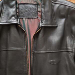 May include: Black leather jacket with a collared neckline and a full-length zipper. The jacket features a small embossed logo on the front and a label inside the collar. The lining is a mix of red, green, and black.