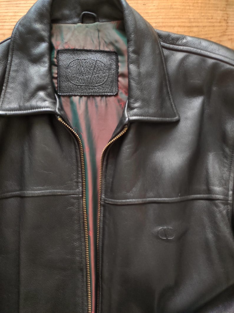 May include: Black leather jacket with a collared neckline and a full-length zipper. The jacket features a small embossed logo on the front and a label inside the collar. The lining is a mix of red, green, and black.