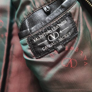 May include: Close-up of a black leather Valentino jacket interior. The label reads "VALENTINO LEATHER CASUAL JACKET 56". The lining is a patterned brown and green fabric. The jacket has a black leather pocket.