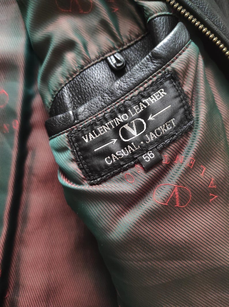 May include: Close-up of a black leather Valentino jacket interior. The label reads "VALENTINO LEATHER CASUAL JACKET 56". The lining is a patterned brown and green fabric. The jacket has a black leather pocket.