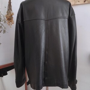 May include: A dark brown leather jacket with a classic collar and long sleeves. The back of the jacket features a yoke detail and vertical seams. The jacket is hanging on a metal stand.
