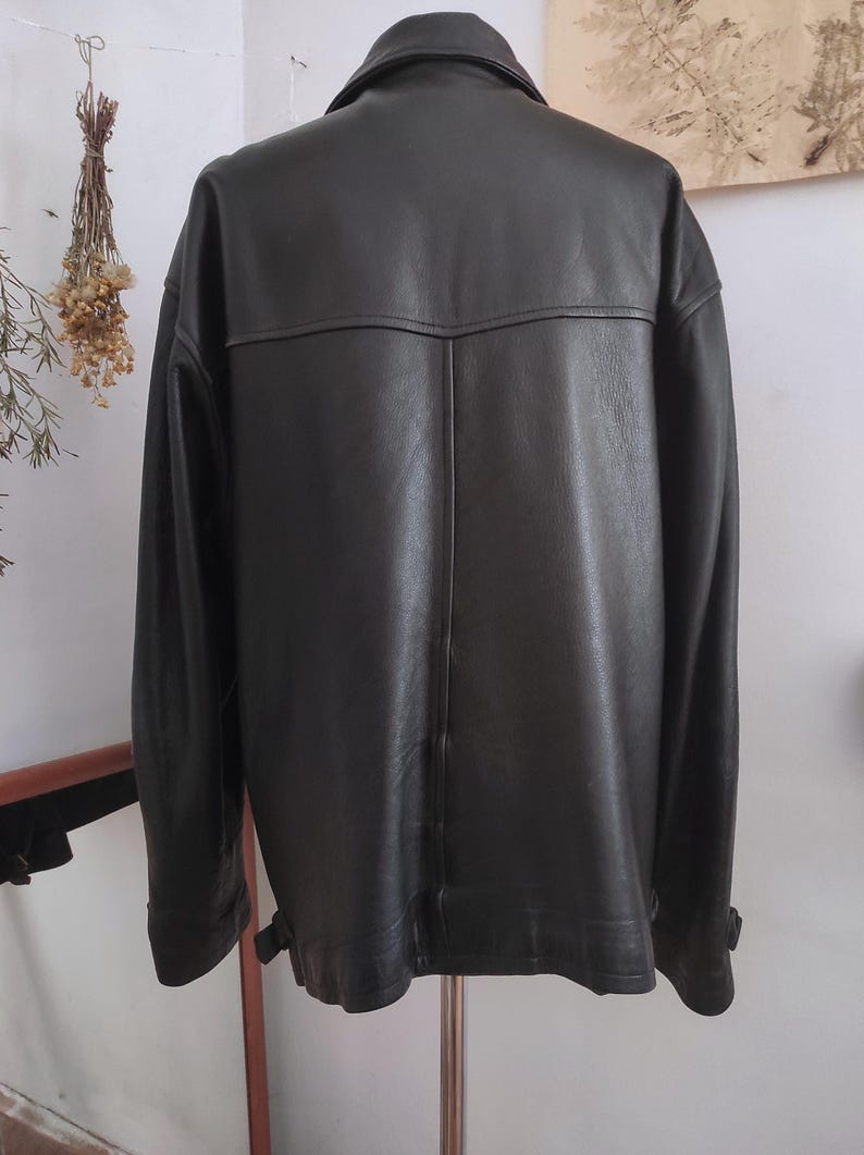 May include: A dark brown leather jacket with a classic collar and long sleeves. The back of the jacket features a yoke detail and vertical seams. The jacket is hanging on a metal stand.