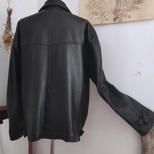 May include: A black leather jacket with a classic design. The jacket has a collared neckline, long sleeves, and a buttoned cuff. The back of the jacket features a yoke detail. The jacket is displayed on a metal stand.