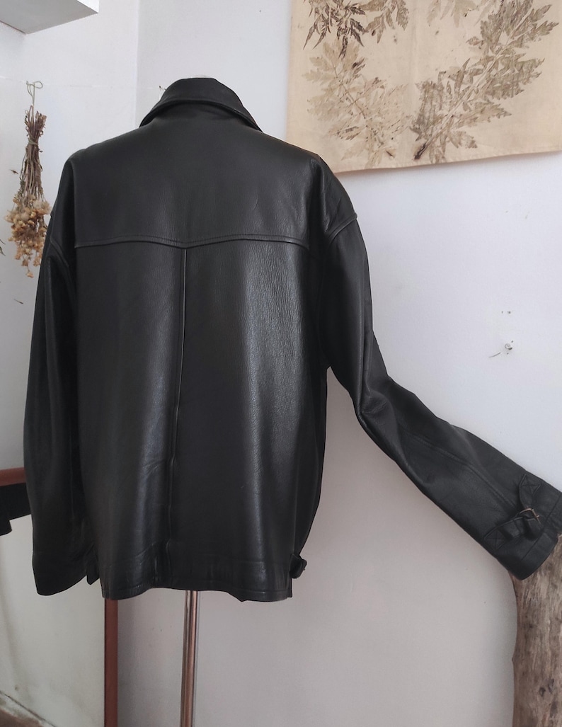 May include: A black leather jacket with a classic design. The jacket has a collared neckline, long sleeves, and a buttoned cuff. The back of the jacket features a yoke detail. The jacket is displayed on a metal stand.